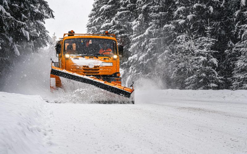 Orange maintenance plough truck on forest road after snowstorm blizzard. Roads get dangerous during winter (driver face blurred)